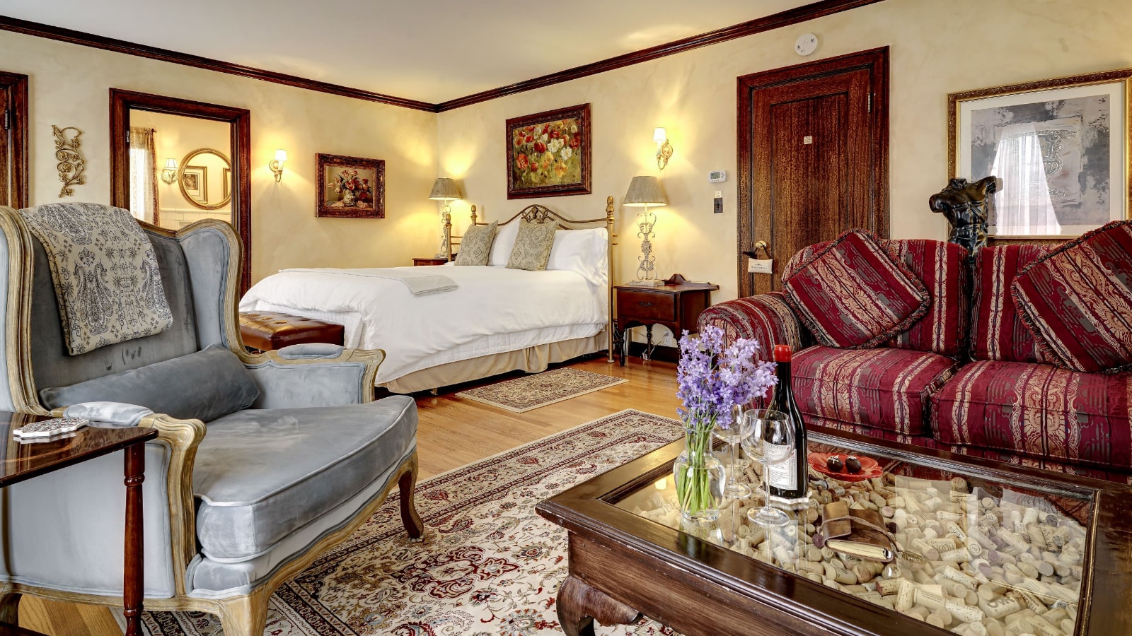 Bedroom with light colored walls, hardwood flooring, area rugs, bed with brass headboard and white linens, red upholstered sofa, gray upholstered chair, and wooden coffee table with glass top