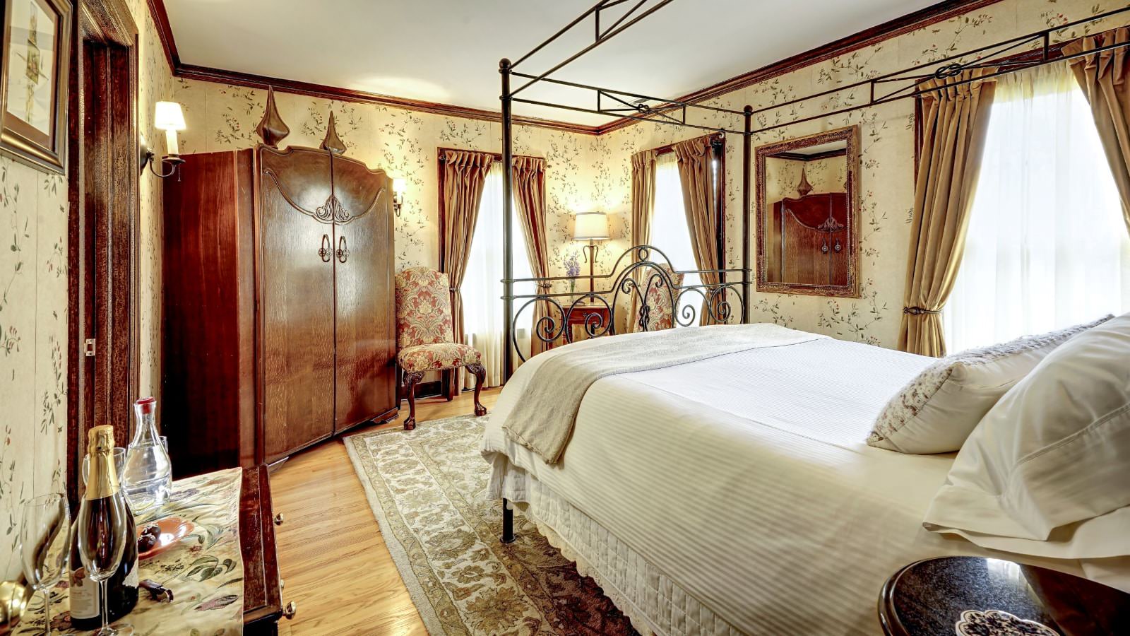 Bedroom with light colored wallpaper, hardwood flooring, area rugs, wrought iron four poster bed with white linens, large wooden armoire, and sitting area