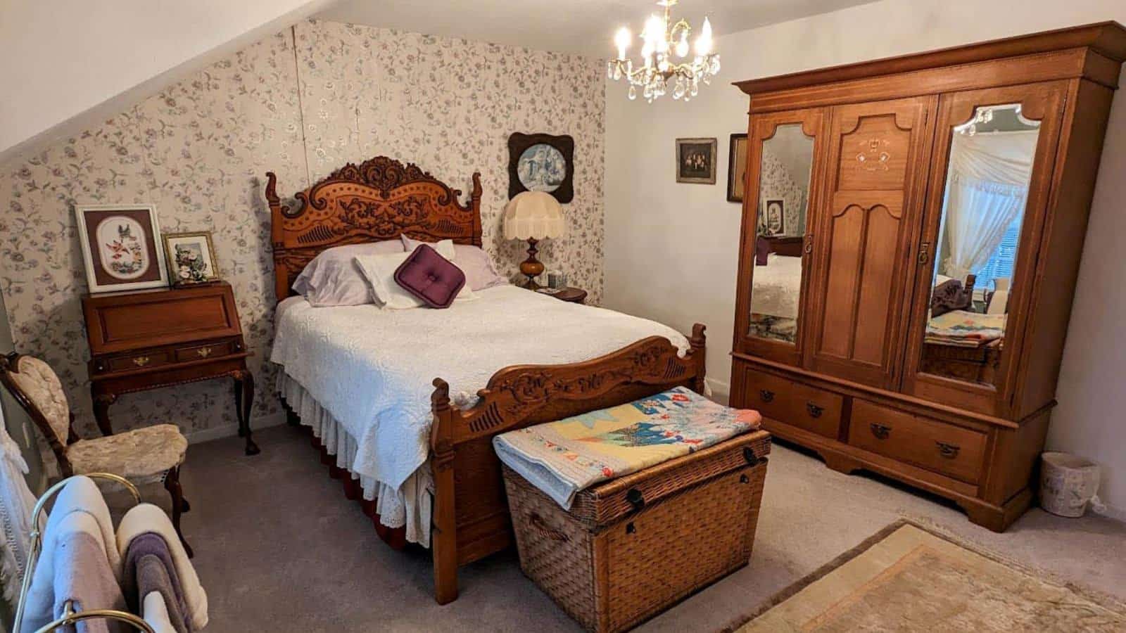 Bedroom with white walls, floral wallpaper, carpeting, wooden antique headboard and foot board with white bedding, and large wooden armoire with mirrors