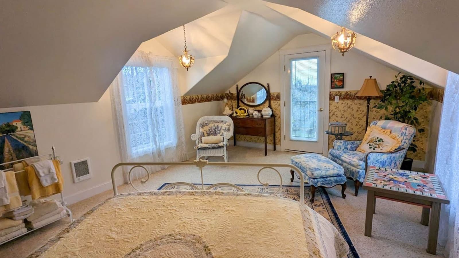 Bedroom with white walls, floral wallpaper, carpeting, white wrought iron bed with light colored quilt, and sitting area