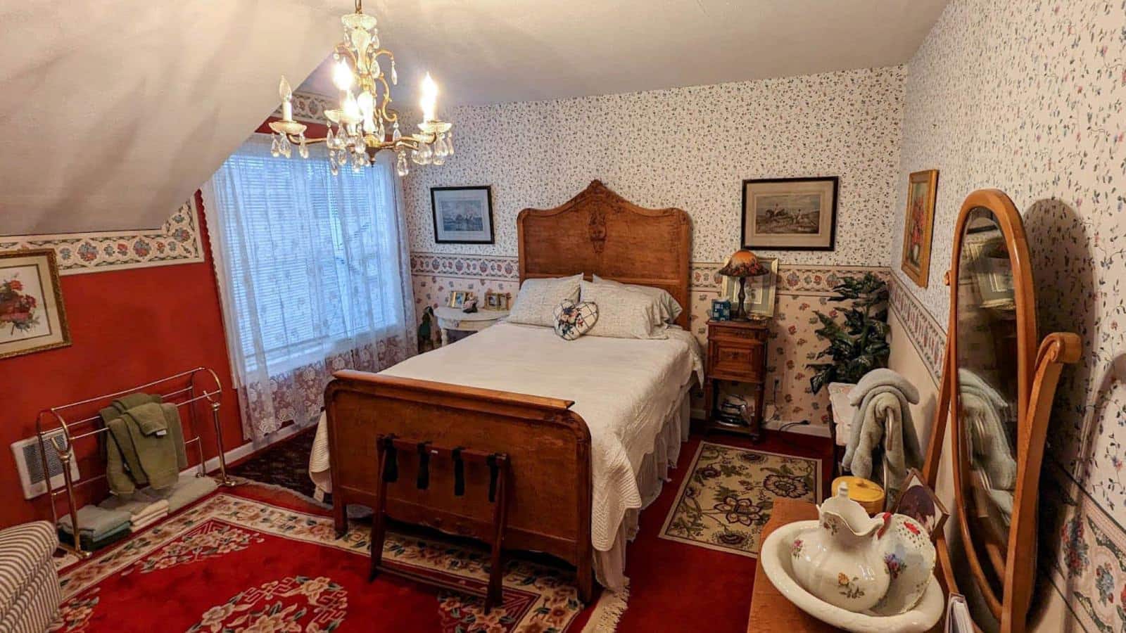 Bedroom with red walls, floral wallpaper, carpeting, wooden antique headboard and foot board with white bedding, and wooden dresser with mirror