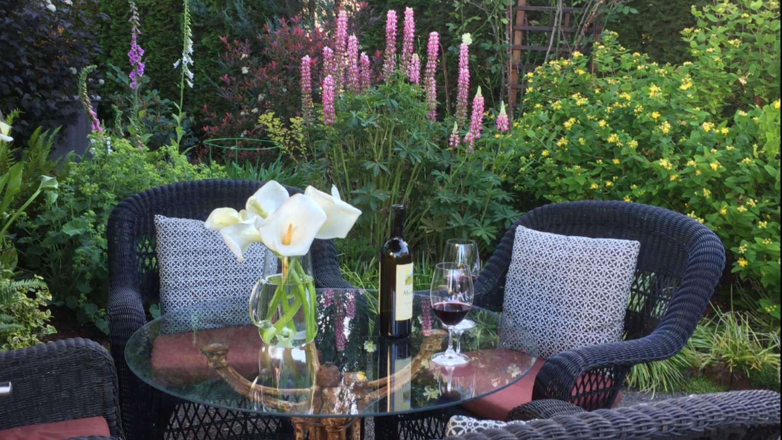 Navy wicker patio chairs and glass table surrounded by lush bushes and flowers