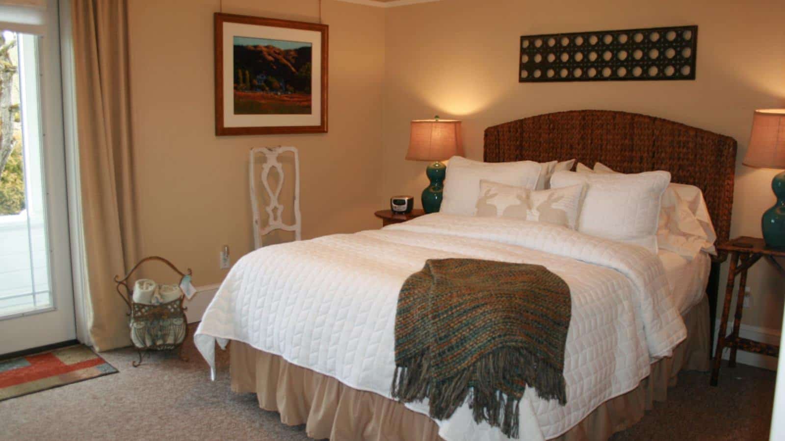 Bedroom with light colored walls, carpeting, brown braided headboard, white bedding, and door to outside