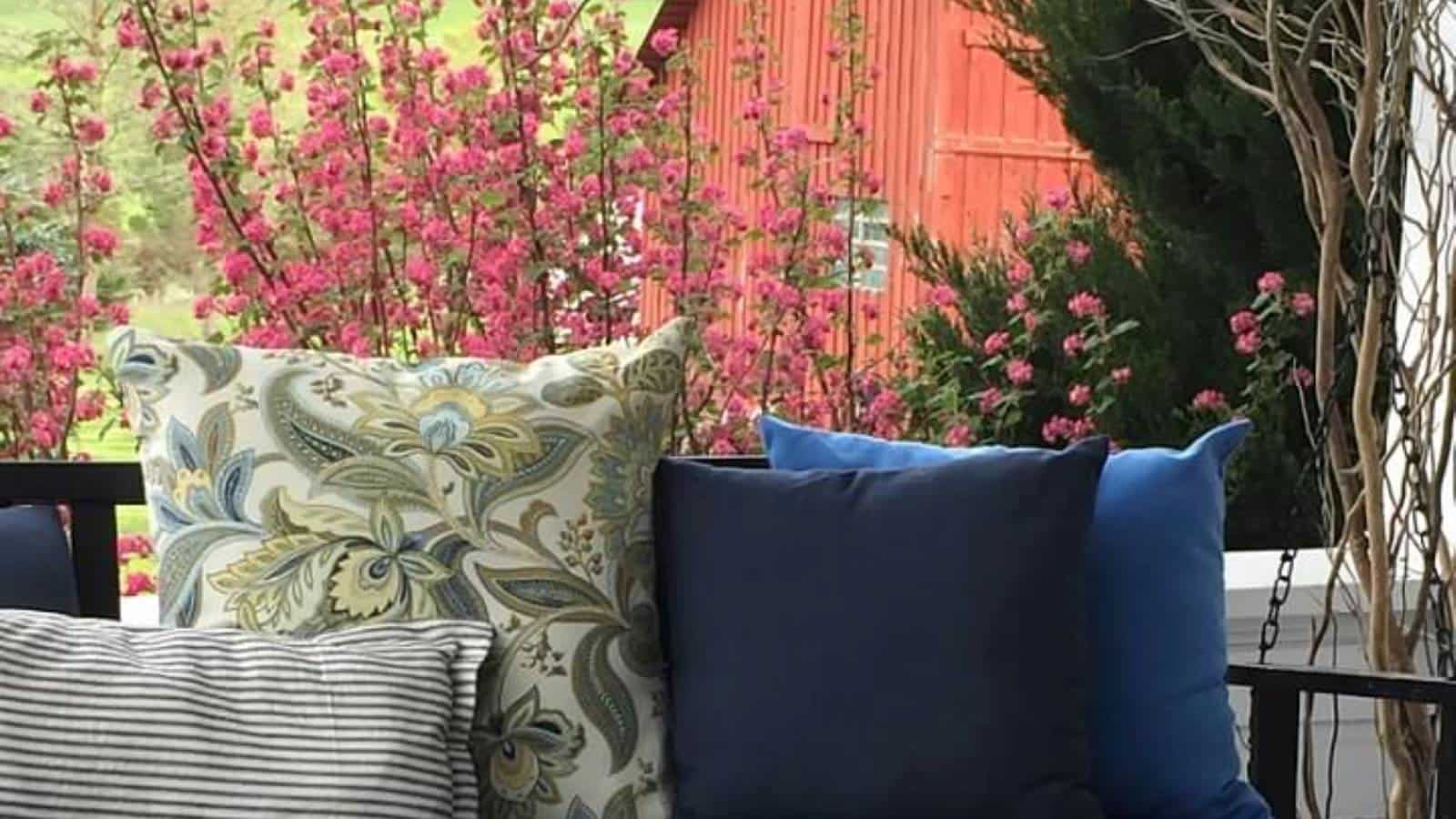 Close up view of porch swing with multicolored pillows surrounded by pink-flowered bushes and red barn in the background
