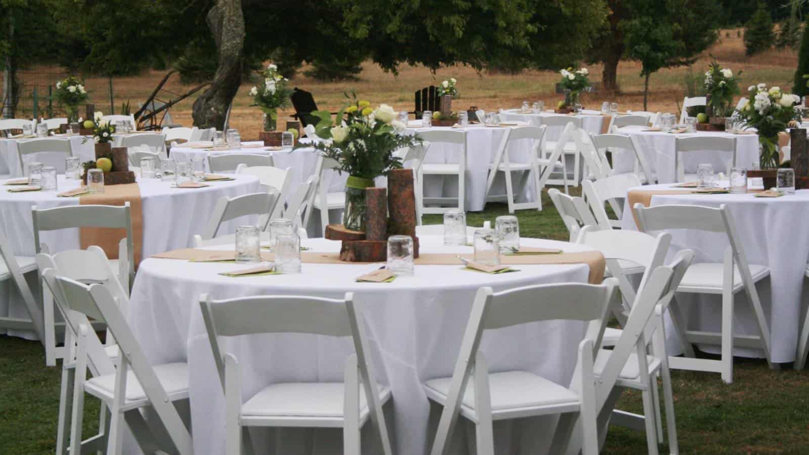 Multiple round tables with white chairs and white tablecloths all set up for an event on green grass next to large green trees