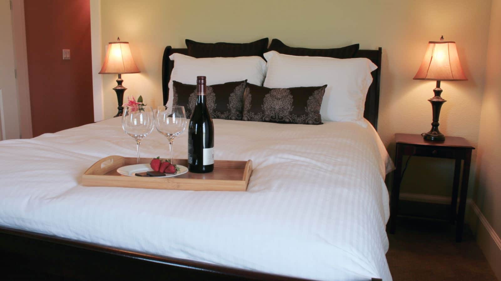 Bedroom with light colored walls, carpeting, dark headboard, white bedding, and wooden tray on bed with fruit, chocolate and wine