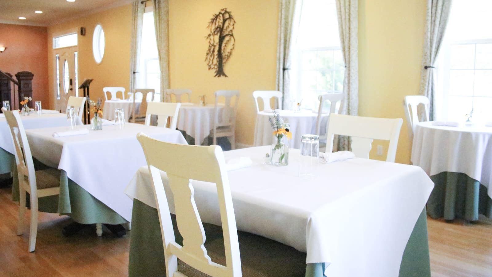 Large dining room with yellow walls, hardwood flooring, and multiple tables with white and green tablecloths and white chairs