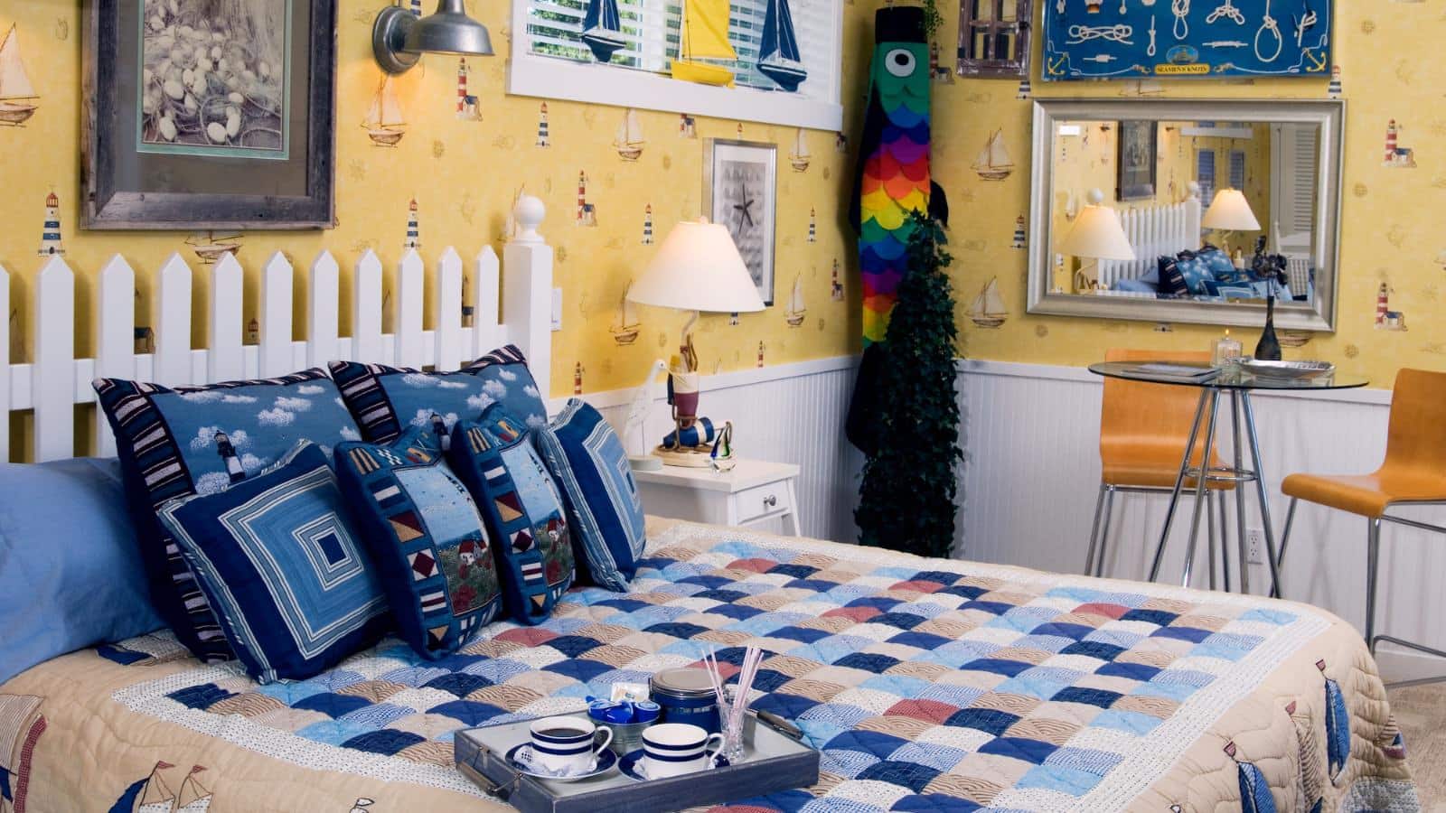 Bedroom with yellow wallpaper with lighthouses, white wainscoting, white picket fence headboard, multicolored bedding, and sitting area