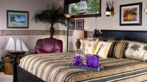 Bedroom with light colored walls and wallpaper, black wooden headboard, multicolored bedding, burgundy leather oversized armchair, and decorative small tree in the corner