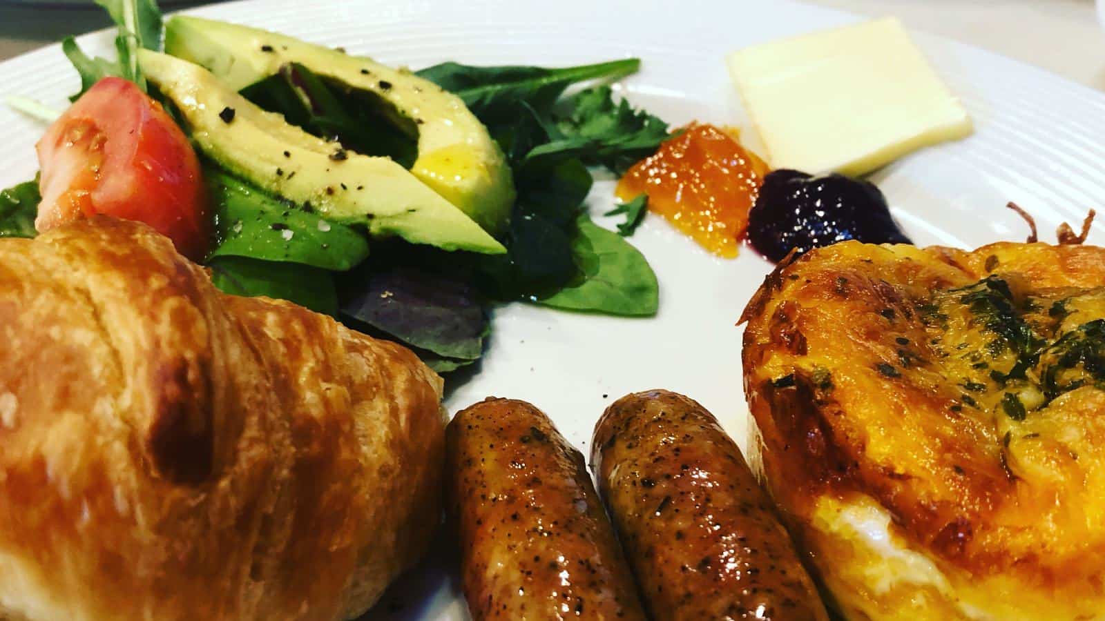 Close up view of white plate with two sausage links, muffin sized quiche, croissant, and sliced avocado and tomato on bed of green spinach