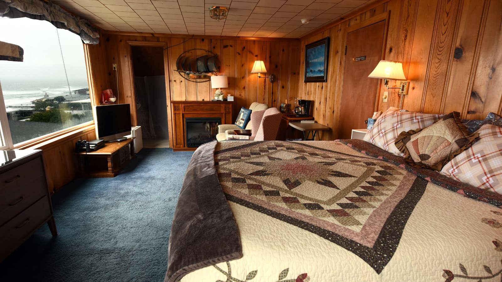 Bedroom with wood paneled walls, carpeting, multicolored bedding, fireplace, and large window with view of the water