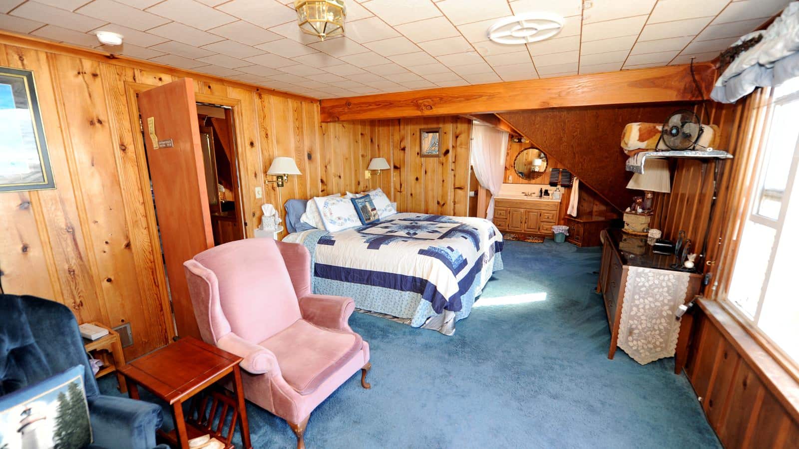 Bedroom with wood paneled walls, carpeting, multicolored bedding, antique pink upholstered armchair, and wooden vanity with sink
