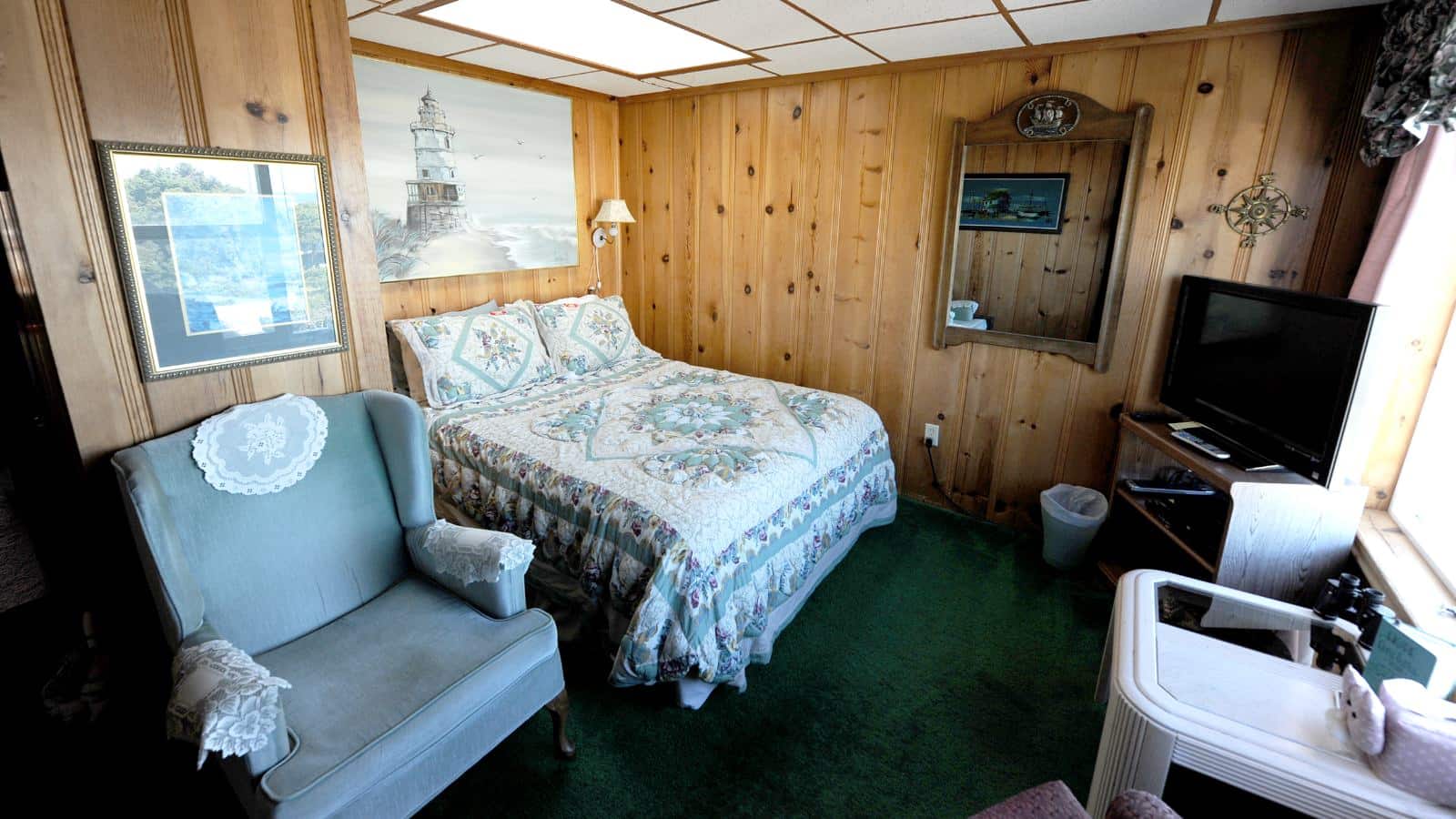 Bedroom with wood paneled walls, carpeting, multicolored bedding, antique blue upholstered armchair, wooden shelving unit with flat panel TV