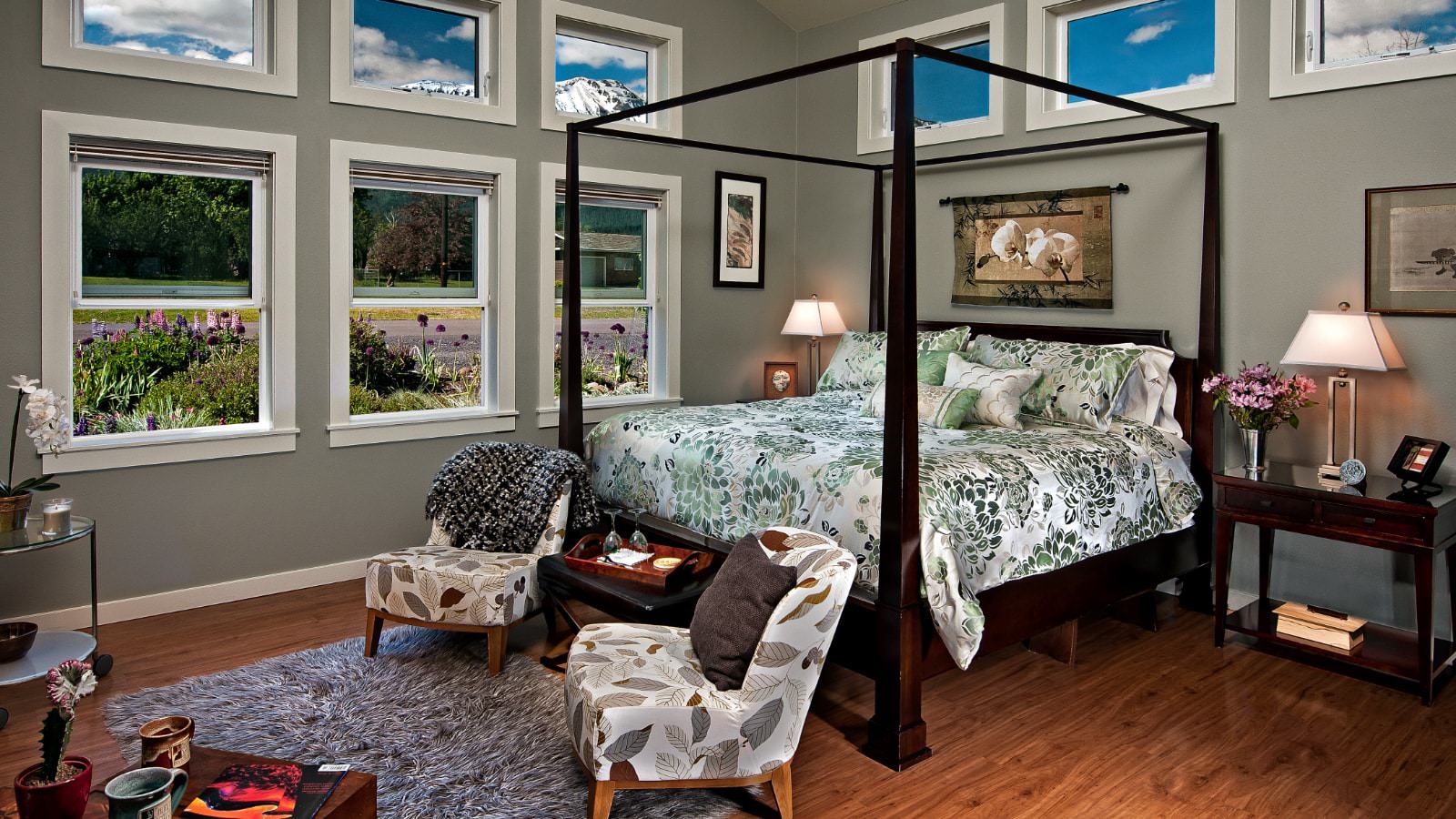 Bedroom with light gray walls, hardwood flooring, vaulted ceiling, four poster dark wooden bed, multicolored bedding, sitting area, and many windows with views of the mountain