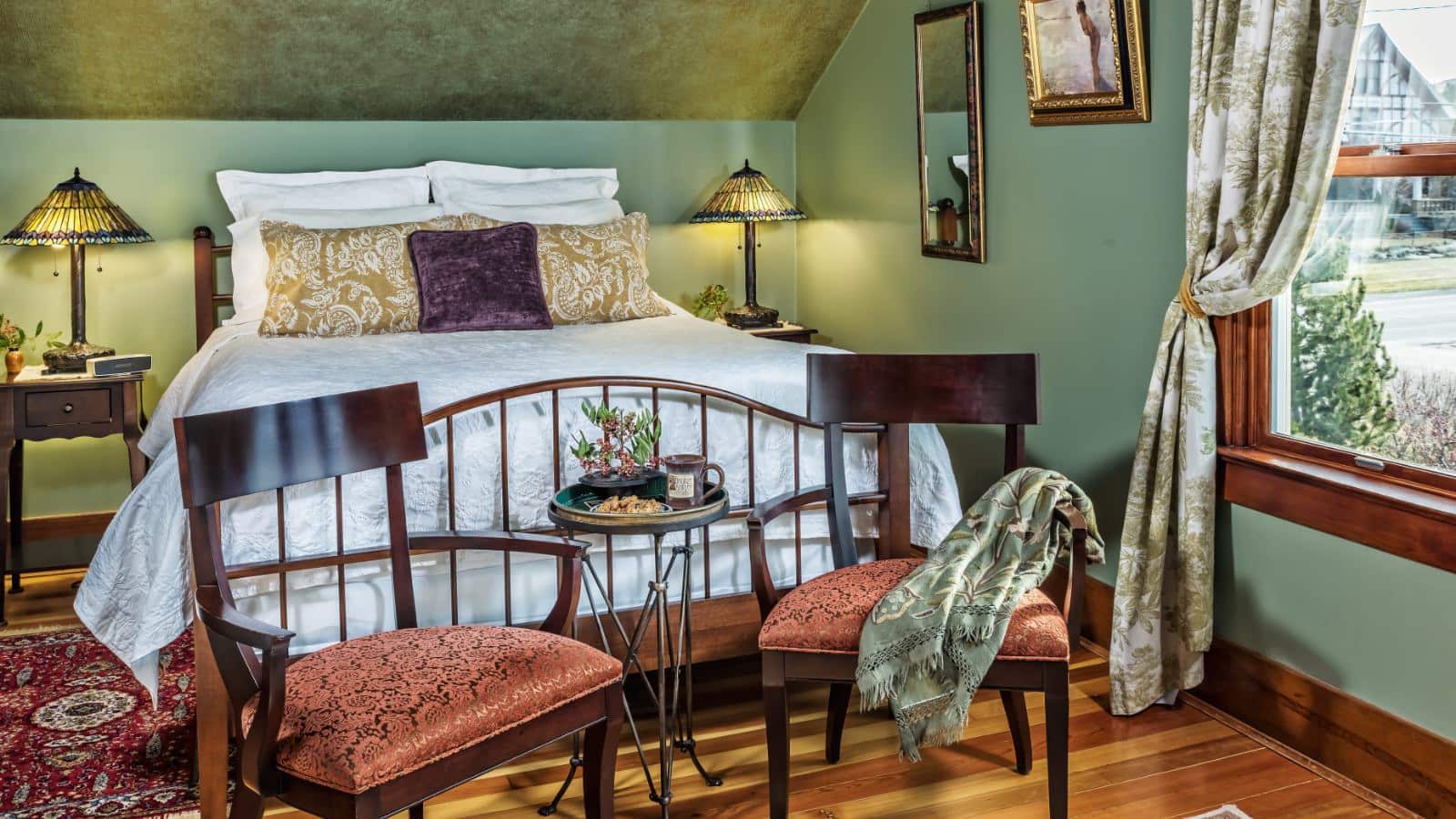 Bedroom with light green walls, hardwood flooring, wooden bed, white bedding, sitting area, and window with views to outside