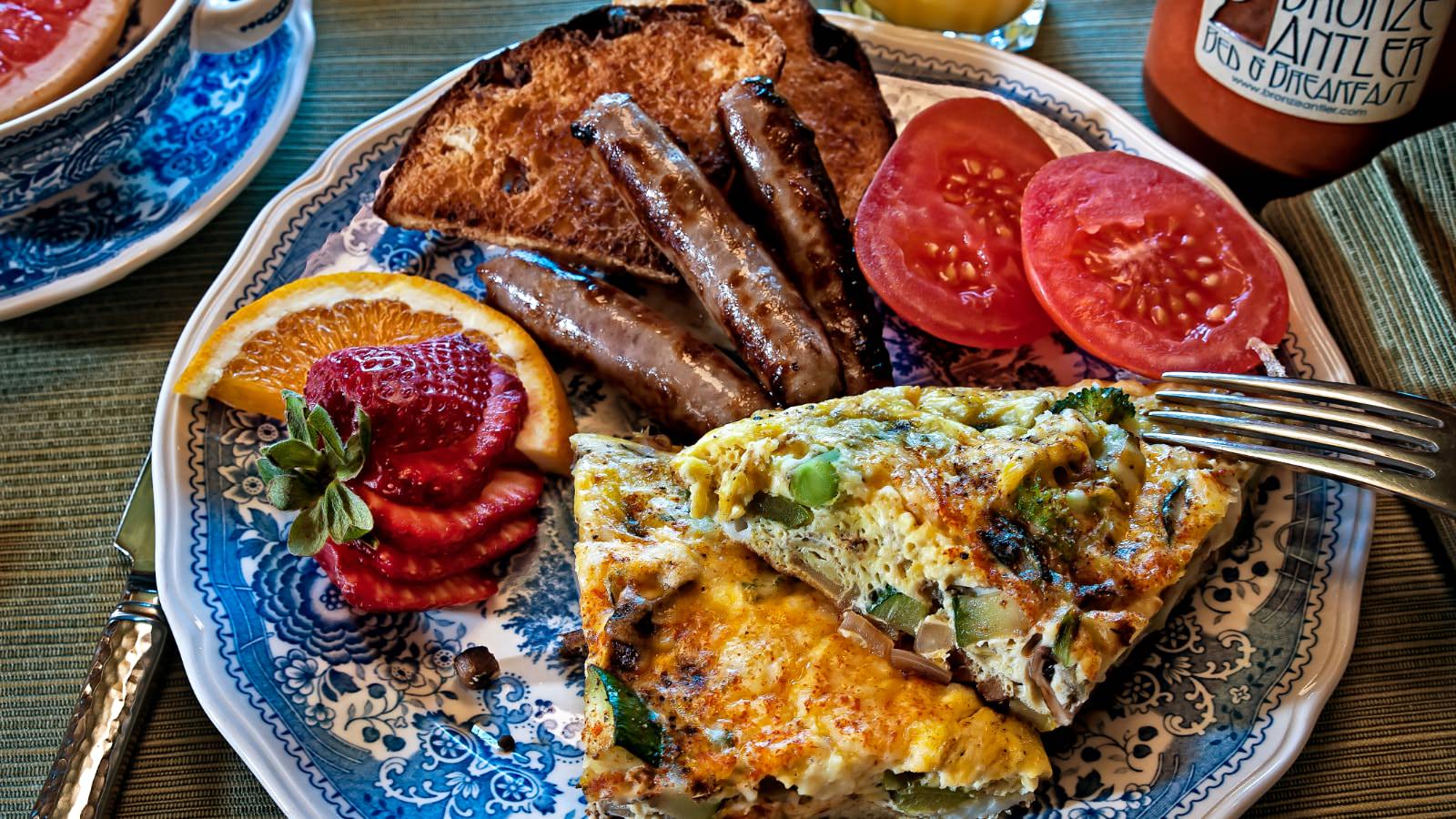 Close up view of blue patterned plate with servings of vegetable quiche, sausage links, toast, and sliced tomatoes, orange, and strawberries