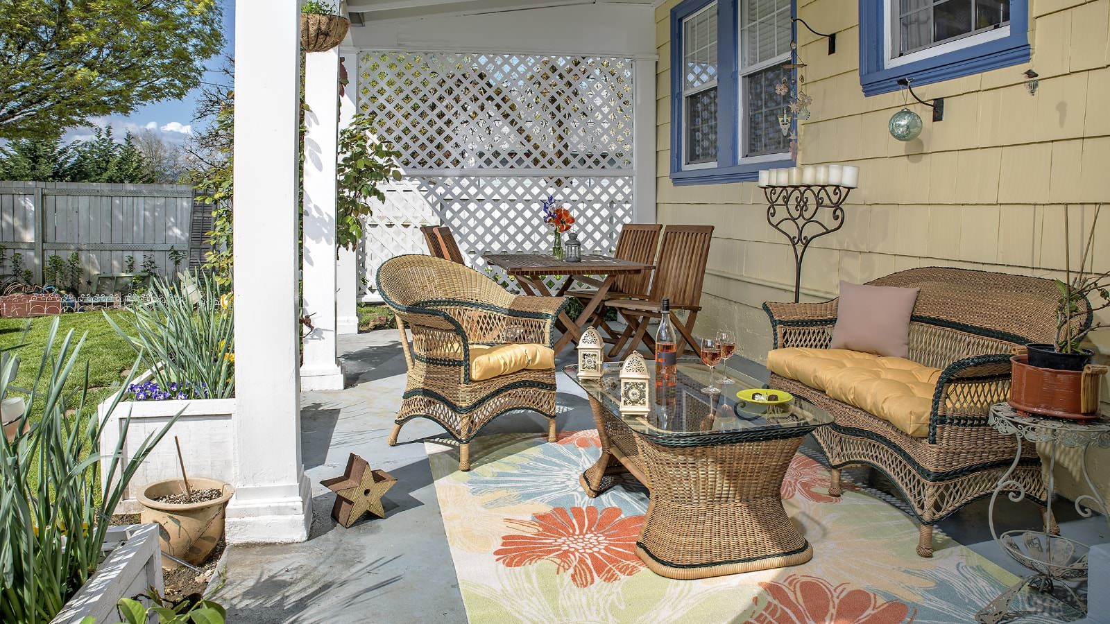 Porch with light colored wicker patio furniture and multicolored area rug surrounded by green grass and flowers