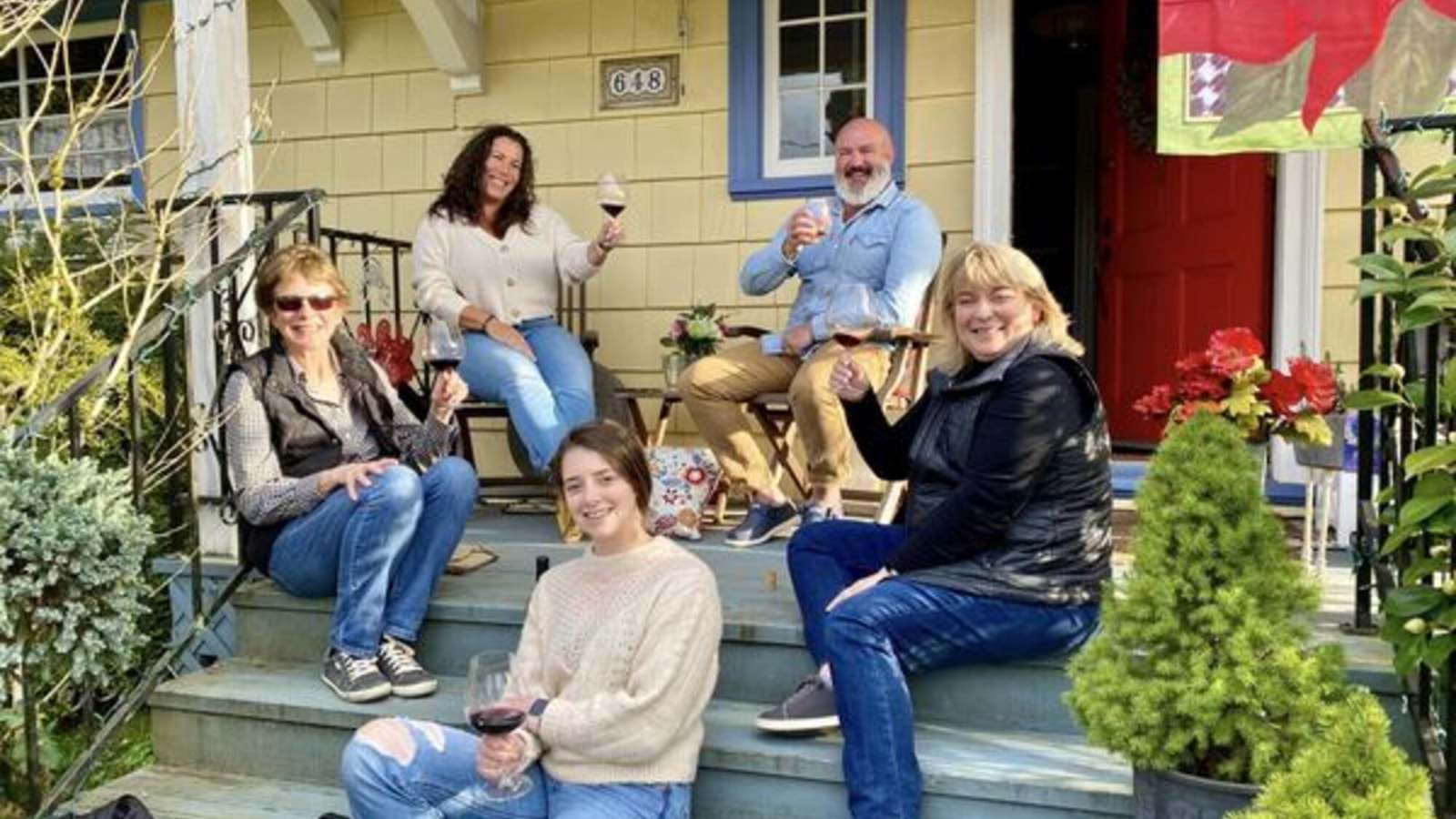 People smiling, holding wine glasses, and sitting on a front porch surrounded by greenery