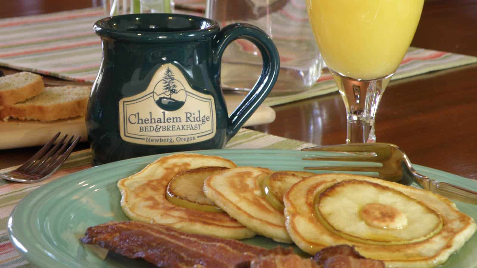 Close up view of light sage green plate with pear pancakes and bacon, glass of orange juice, and coffee cup with Chehalem Ridge Bed & Breakfast text and logo