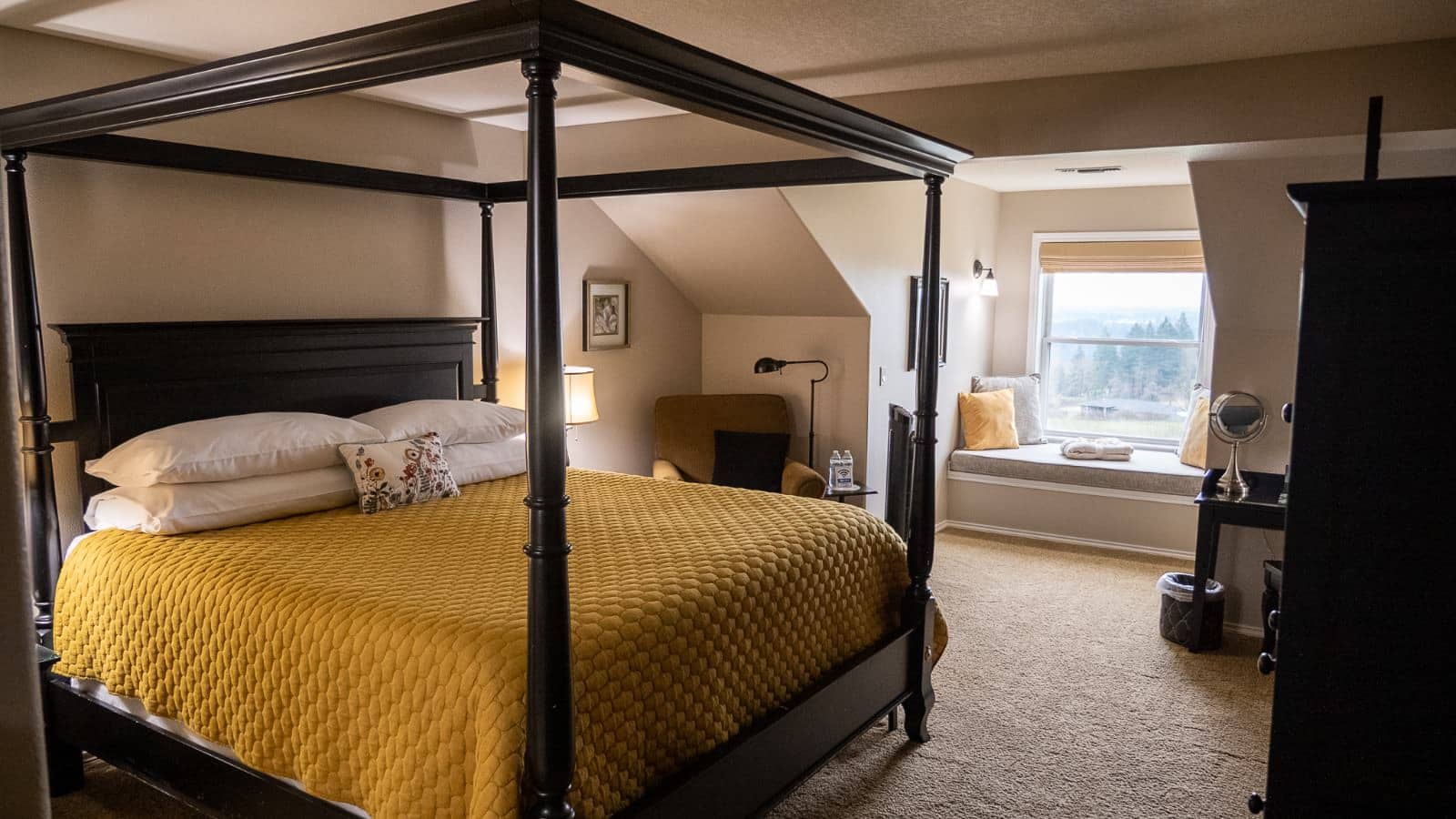 Bedroom with light tan walls, carpeting, dark wooden four poster bed with white pillows and mustard comforter, dark wooden dresser, and nook with views of the outside