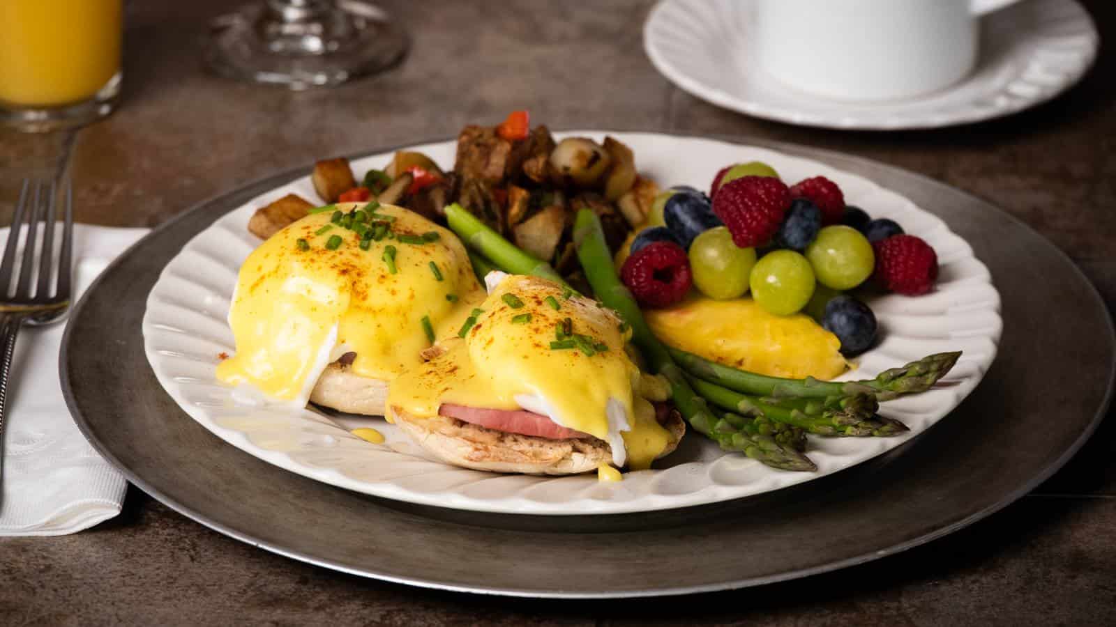 Close up view of white plate with eggs benedict, home fries, asparagus, and fruit