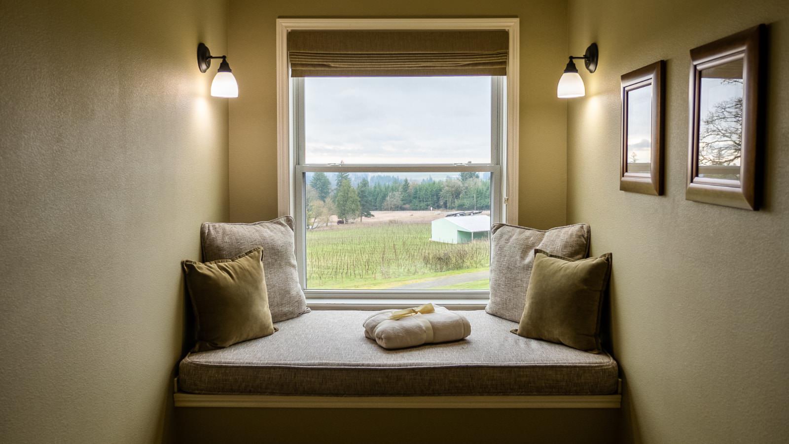 Nook with light tan walls, two wall sconces, tan cushion, tan and green pillows, and window with view of a vineyard