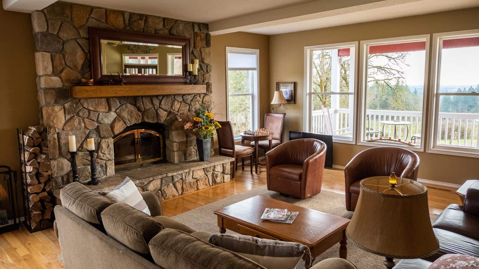 Living room with light tan walls, white trim, hardwood flooring, large stone fireplace, tan upholstered sofa, two leather chairs, and large windows with views of the outside