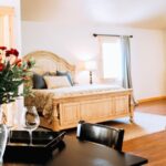 Large bedroom with light colored walls, hardwood flooring, vaulted ceiling, light wooden bed, upholstered sofa, and black table and chairs
