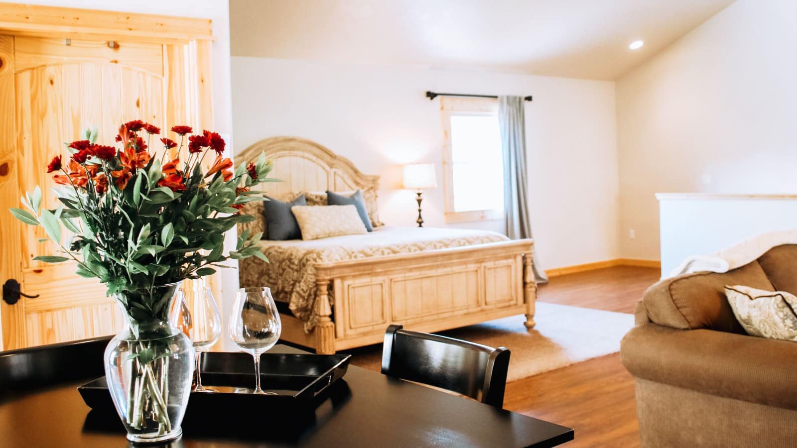 Large bedroom with light colored walls, hardwood flooring, vaulted ceiling, light wooden bed, upholstered sofa, and black table and chairs