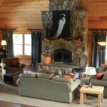 Large great room with wood paneling on the walls, hardwood flooring, upholstered sofa, loveseat and armchairs, large stone fireplace, and black piano in the corner