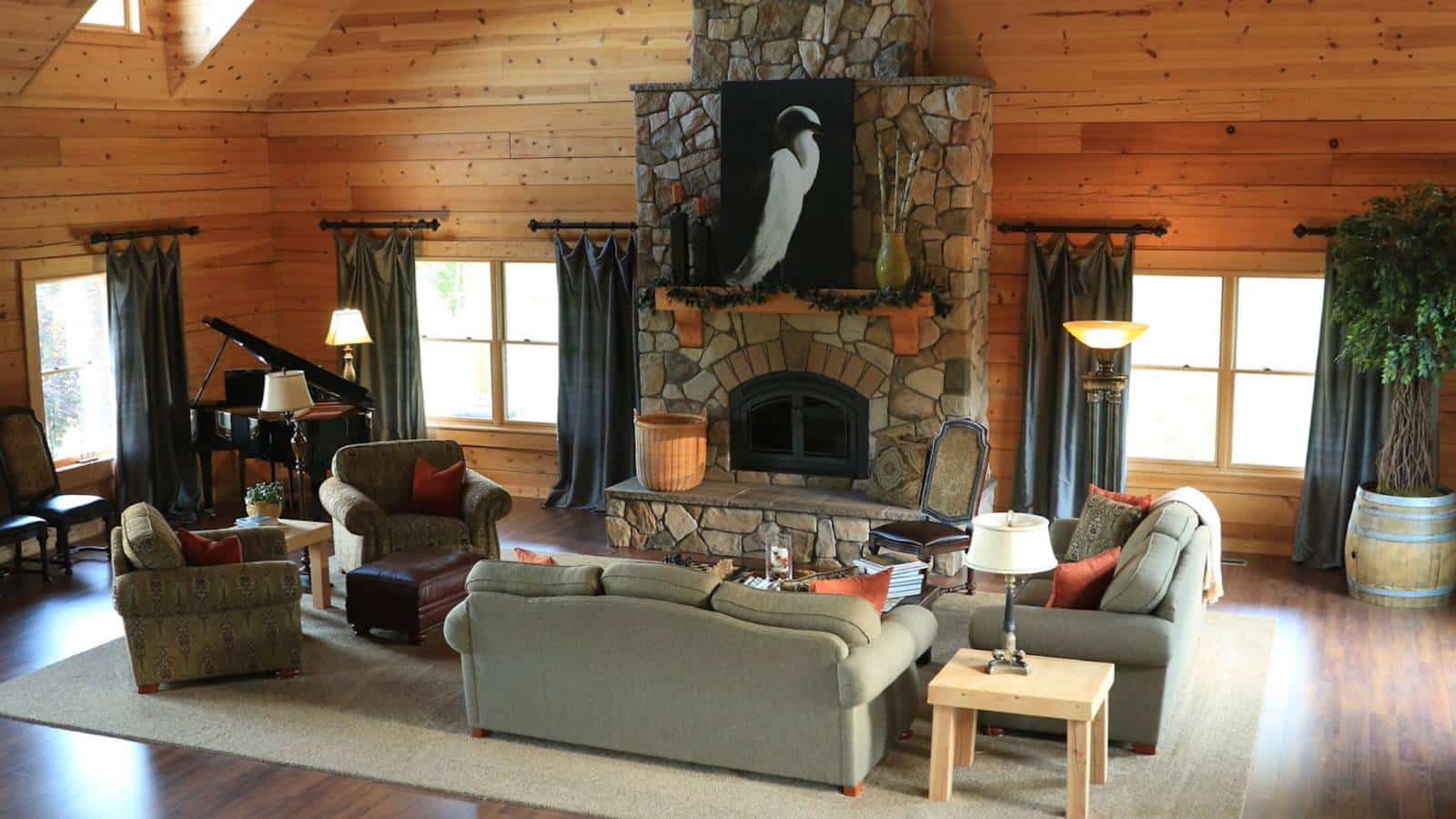 Large great room with wood paneling on the walls, hardwood flooring, upholstered sofa, loveseat and armchairs, large stone fireplace, and black piano in the corner