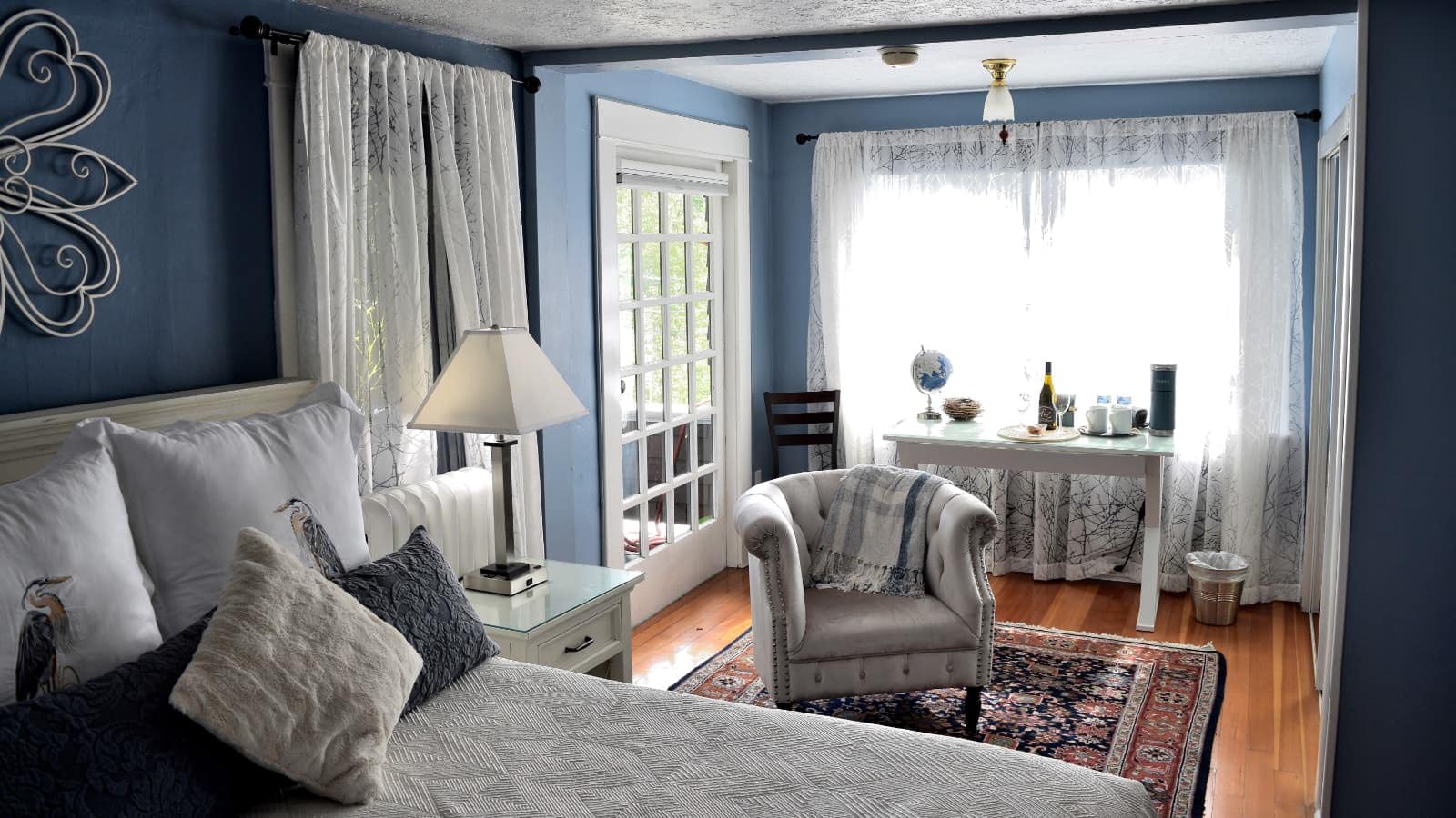Bedroom with blue walls, hardwood flooring, white headboard, white bedding, antique upholstered armchair, and large windows