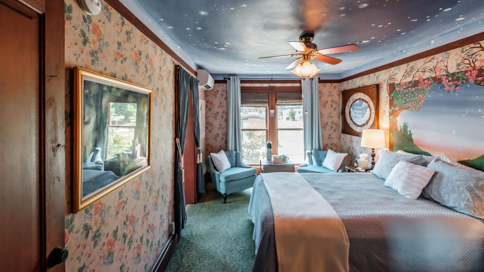 Bedroom with floral wallpaper, painted scene on wall above bed, light blue bedding, and sitting area
