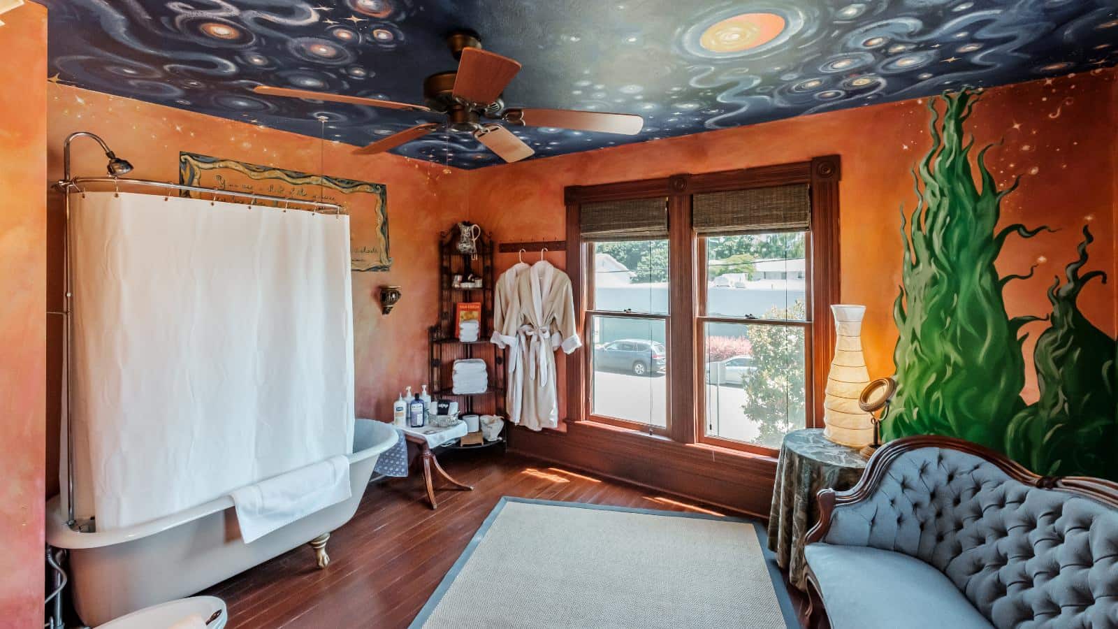Bedroom with custom scenes painted on the walls and ceiling, hardwood flooring, claw-foot tub with shower, and antique wooden and blue upholstered love seat