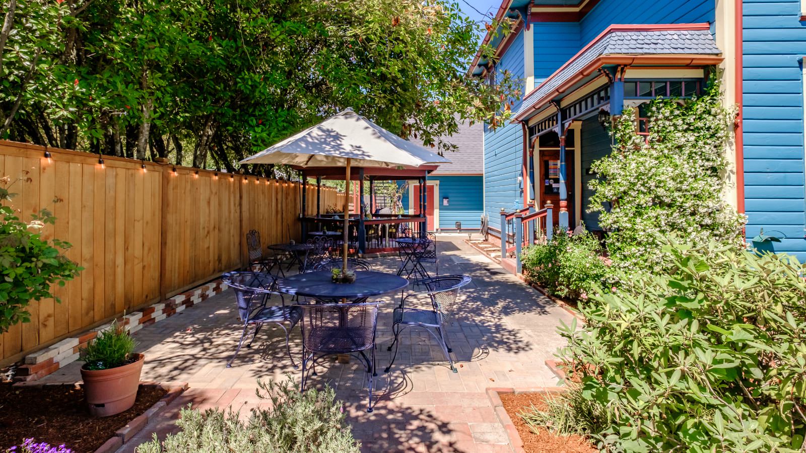 Exterior light stone brick patio with wrought iron patio furniture, umbrella, green bushes, and wooden fence lined with Edison lights