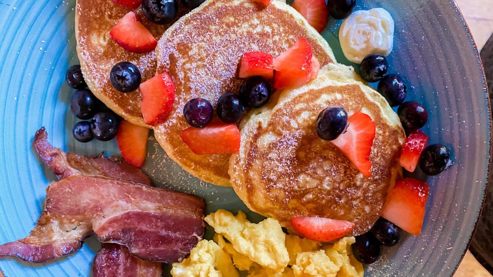 Close up view of blue plate with scrambled eggs, bacon, and pancakes topped with strawberries, blueberries, and powdered sugar