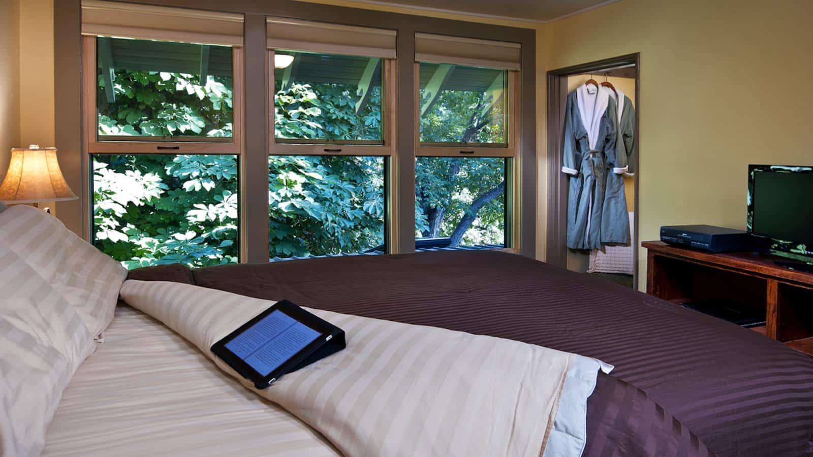 Bedroom with light green walls, multicolored bedding, and large windows with view of green trees