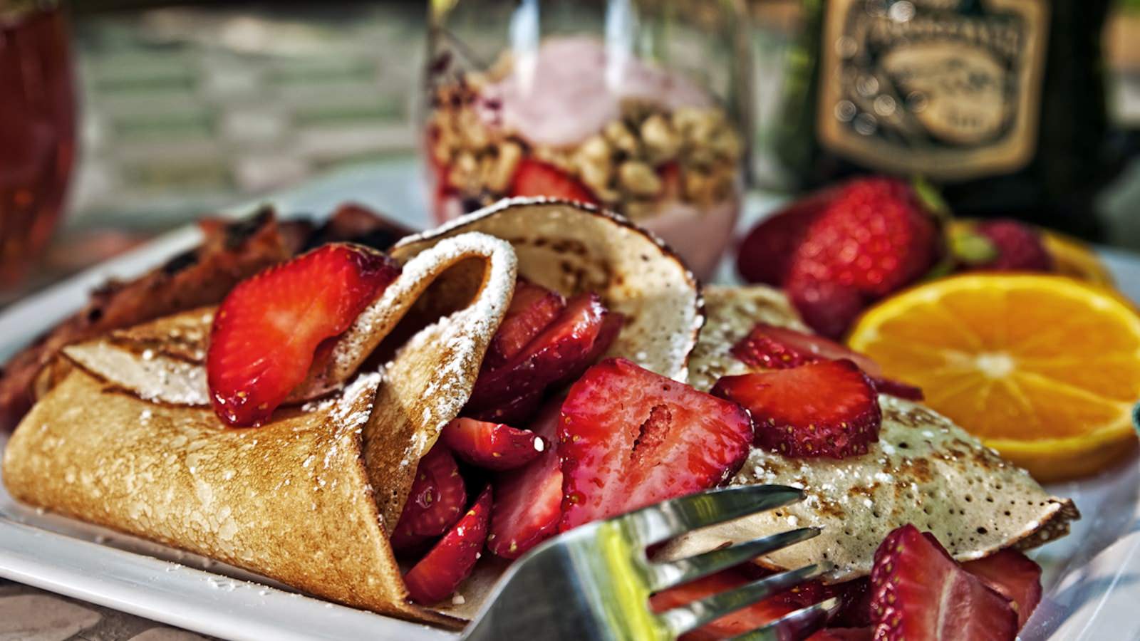 Close up view of white plate with crepes covered with strawberries, sliced orange, and parfait cup