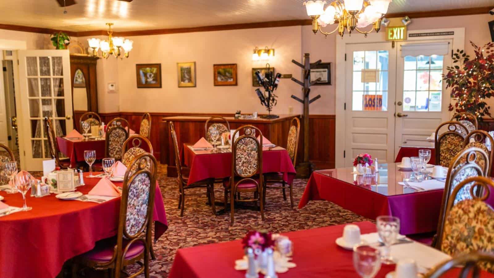 Dining room with light colored walls and wooden wainscoting, carpeting, and many tables with red tablecloths and wooden chairs