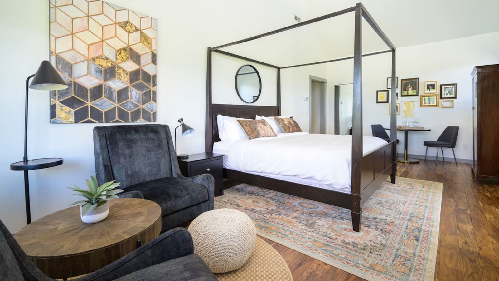 Bedroom with white walls, hardwood flooring, dark wooden four poster bed, white bedding, dark gray upholstered chairs and a sitting area