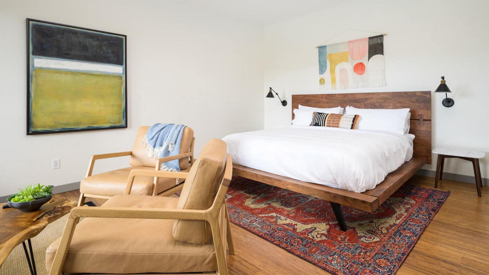 Bedroom with white walls, hardwood flooring, brown platform bed, white bedding, and light tan leather chairs