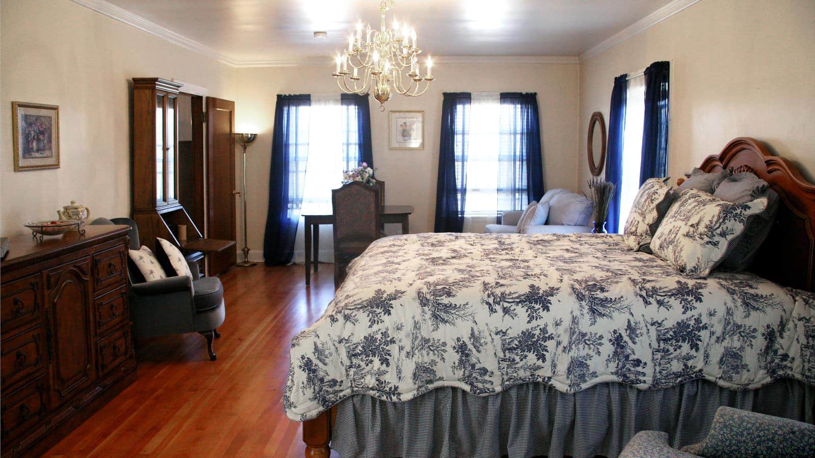 Bedroom with cream walls, hardwood flooring, wooden bed, multicolored bedding, dark wooden dresser, and many windows with navy blue sheer curtains