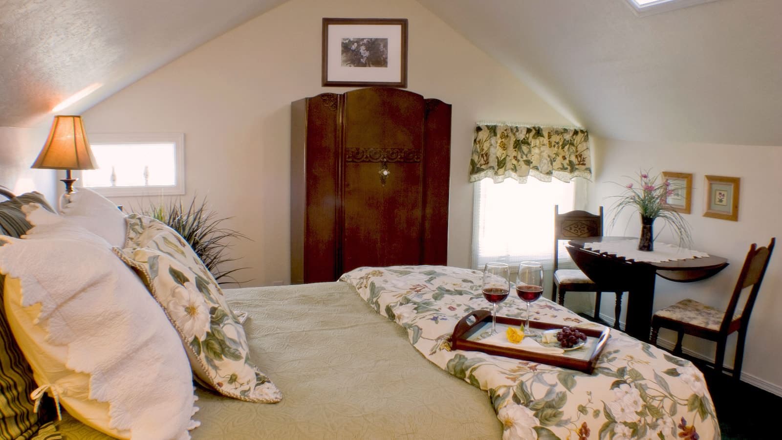 Bedroom with white walls, carpeting, multicolored floral bedding, dark wooden antique armoire, and sitting area