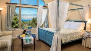 Bedroom with light colored walls, carpeting, blue wooden four poster bed with canopy curtains, multicolored wedding, upholstered loveseat, and large window with views of the water