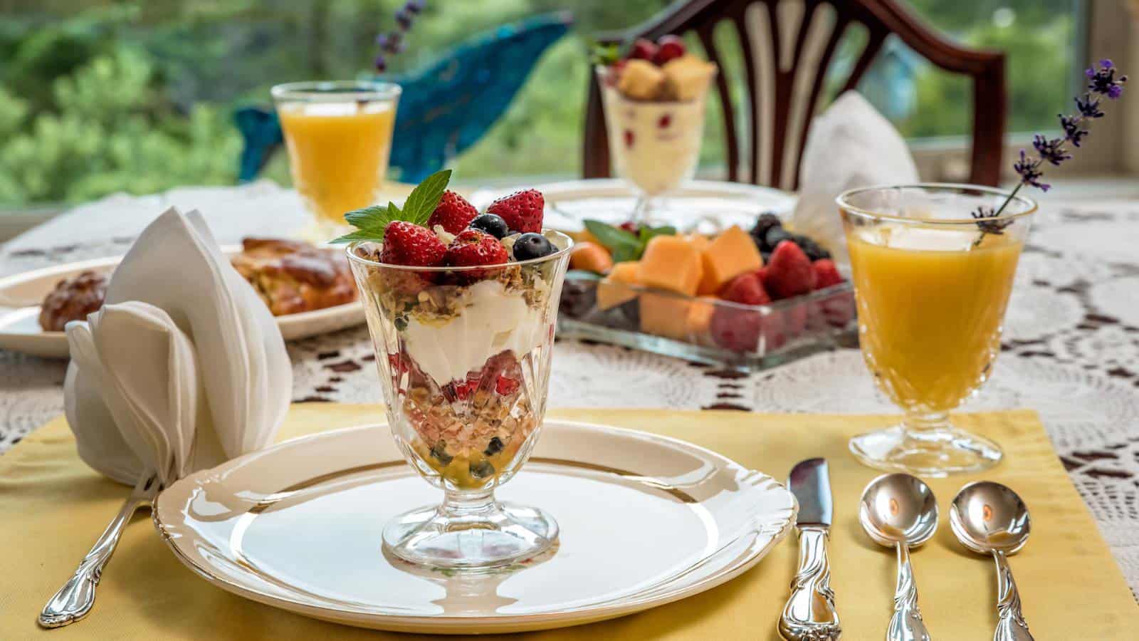Close up view of fruit parfait in clear glass cup sitting on white plate with other breakfast dishes in the background