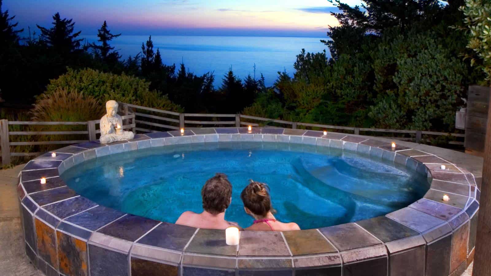 Man and woman sitting in large tiled hot tub looking out to the ocean at dusk