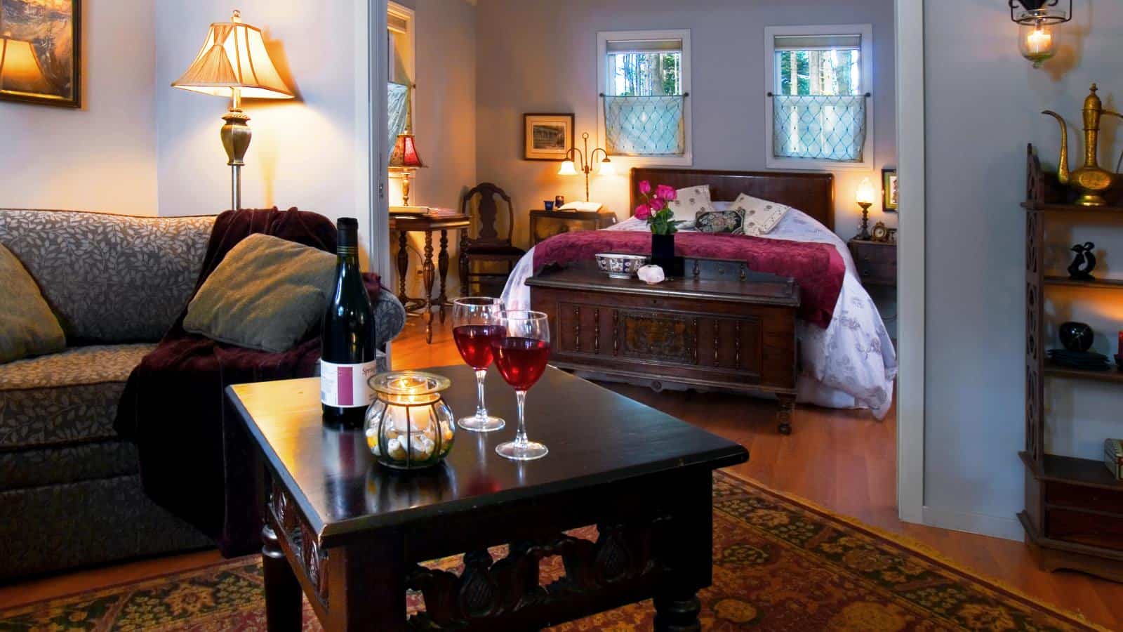 Large bedroom suite with light colored walls, hardwood flooring, upholstered sofa, wooden coffee table, wooden bed, purple bedding, and large wooden trunk at foot of bed