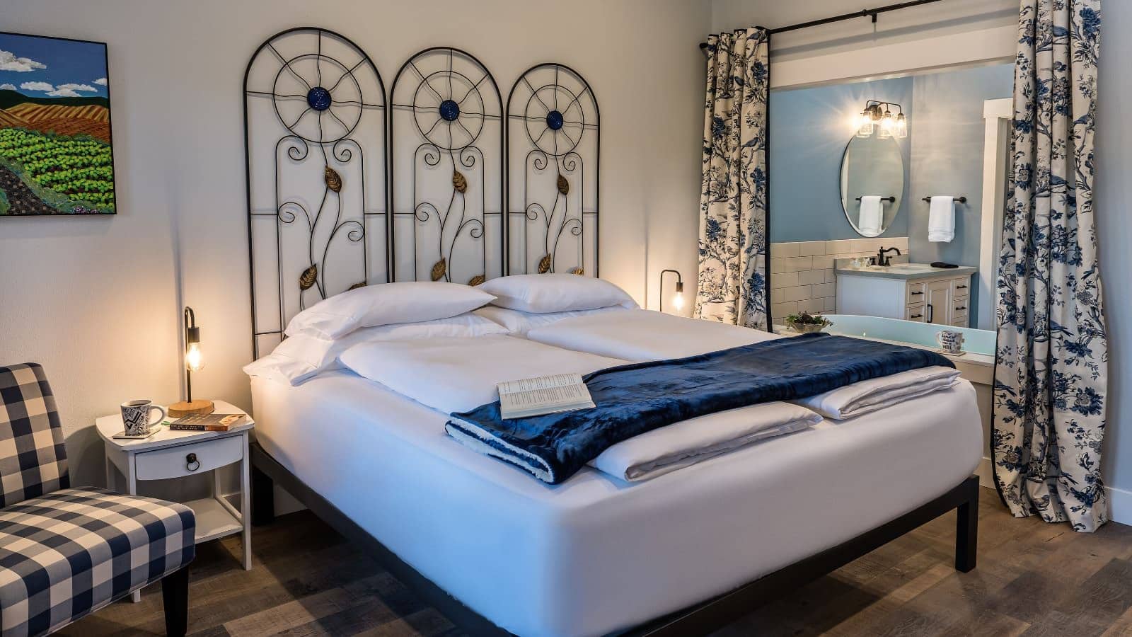 Bedroom with light colored walls, hardwood flooring, custom metal headboard, white bedding, navy and white checked upholstered chair, and view into the bathroom