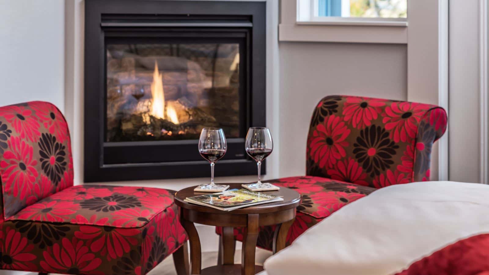 Close up view of two glasses of red wine on a small wooden table near two upholstered chairs with a red and black flower print and fireplace in the background