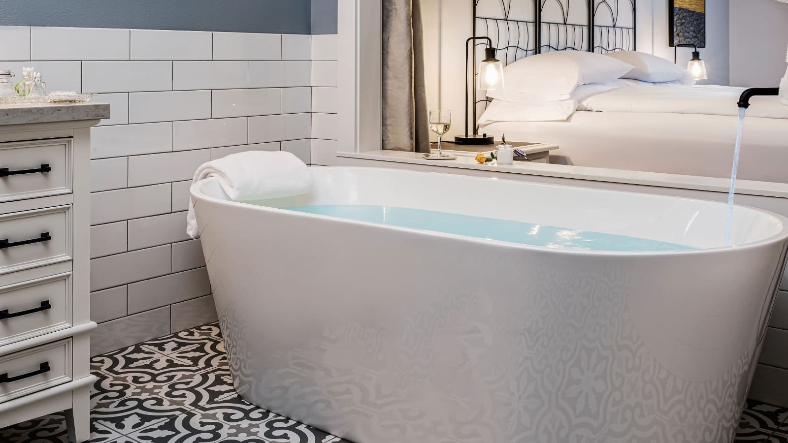 Bathroom with white tiled wainscoting, filigree floor tiles, and large white bathtub full of water